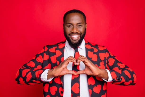 A smiling man in a heart-patterned suit making a hand heart, representing the traditional, surface-level charm that often masks a lack of genuine compatibility on first dates.
