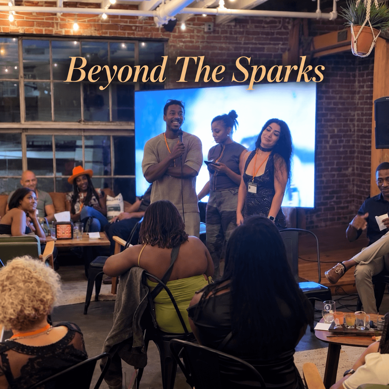 Beyond The Sparks host speaks into a microphone beside two participants while seated guests watch in a brick-walled event space, Attractive People